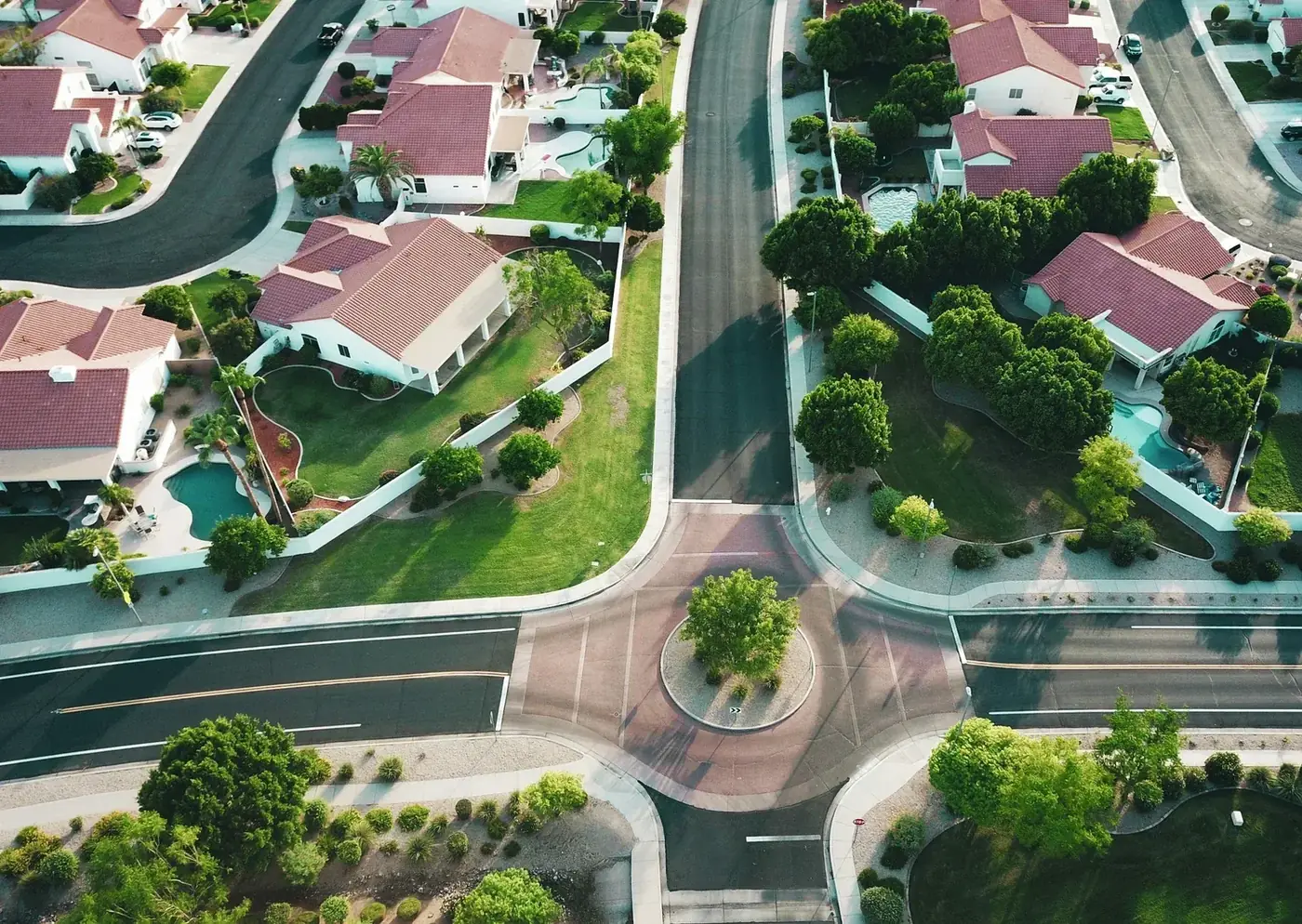 Aerial view of suburban neighborhood with houses, roads, green lawns, and trees, highlighting a roundabout in the center.